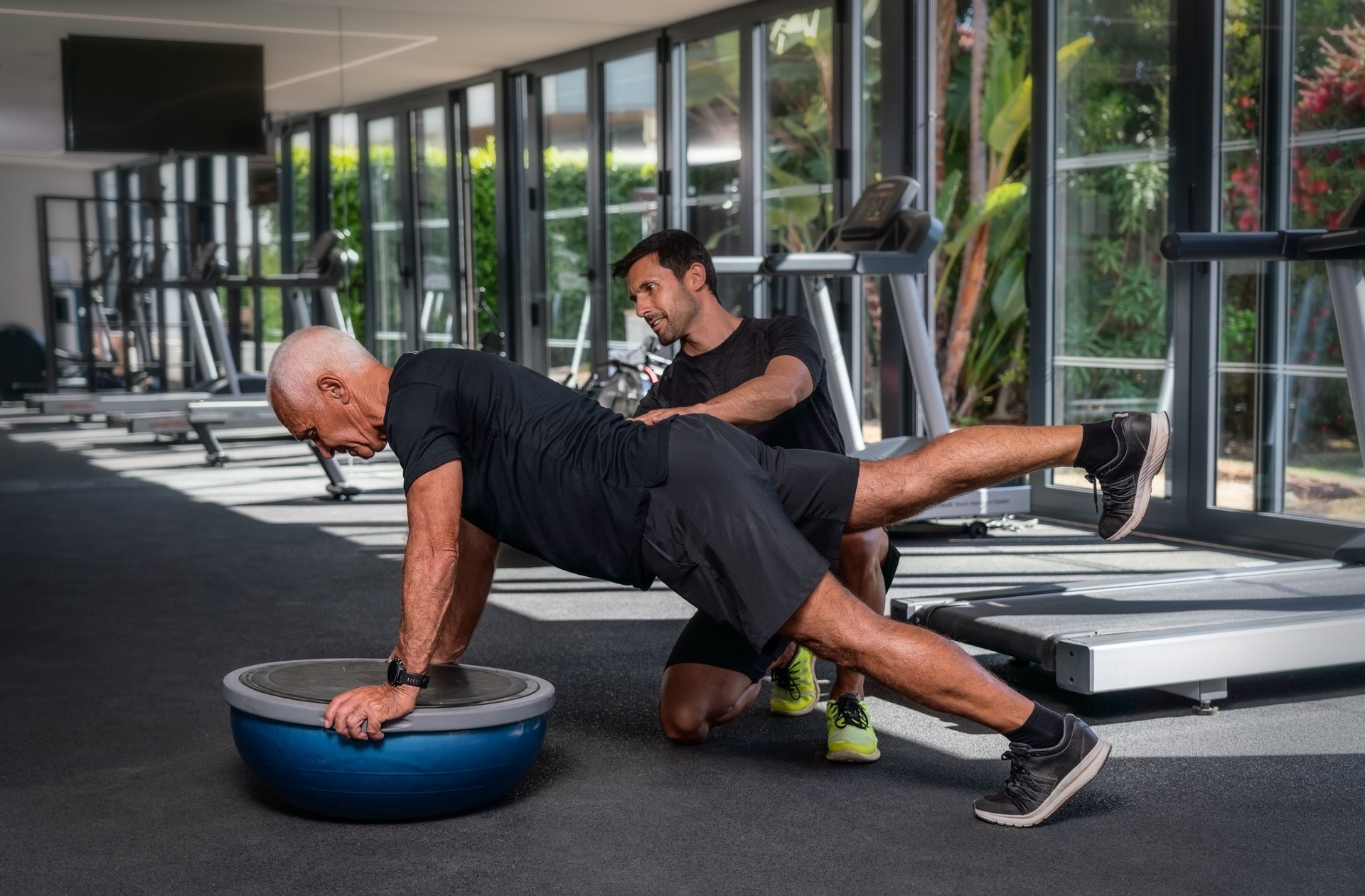 An elderly Caucasian man, in the gym, with a personal fitness trainer, in rehabilitation, doing exercises for balance on a gymnastic ball.